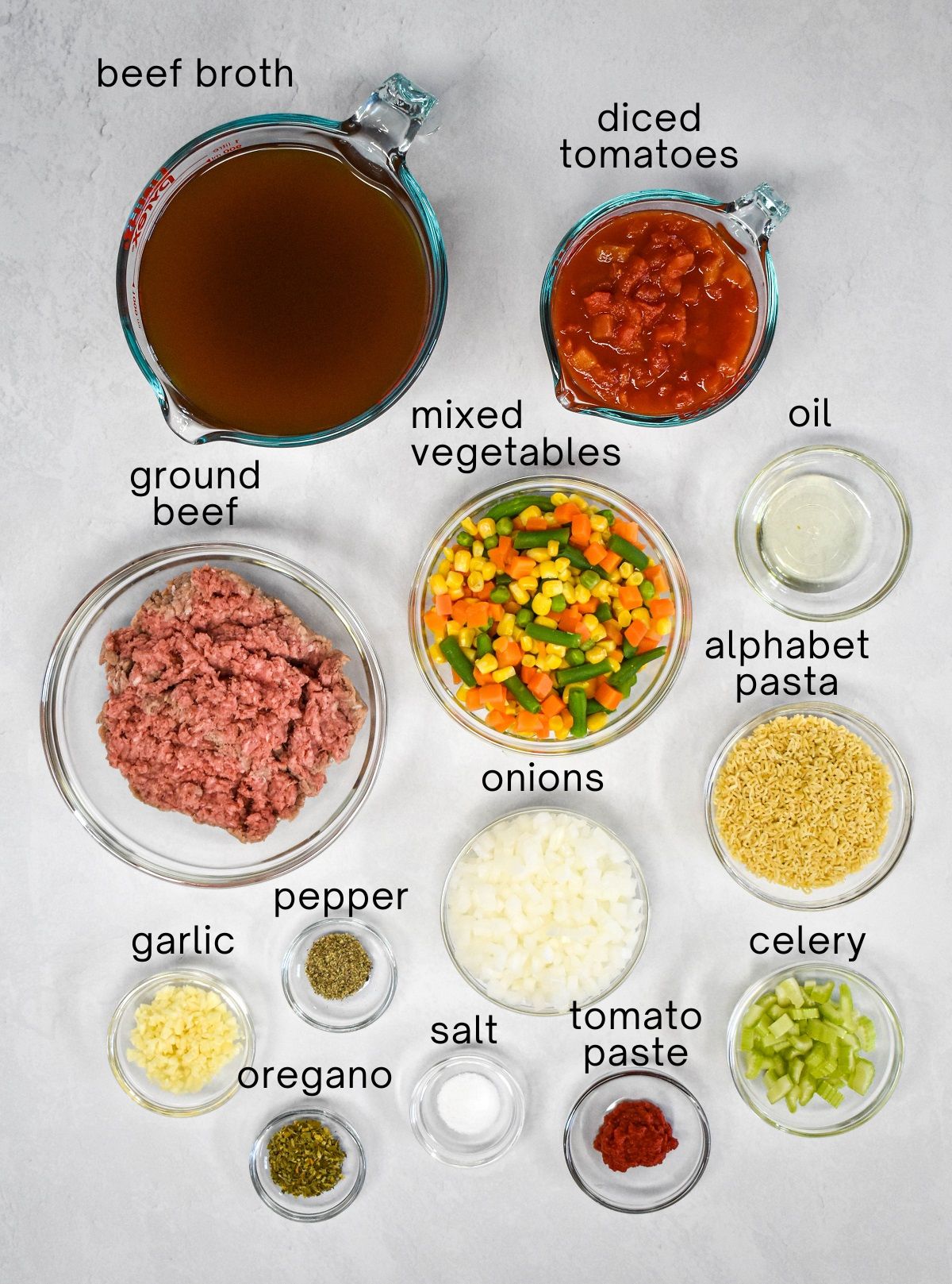 The ingredients for the soup prepped and arranged in glass bowls on a white table with each labeled with small, black letters.