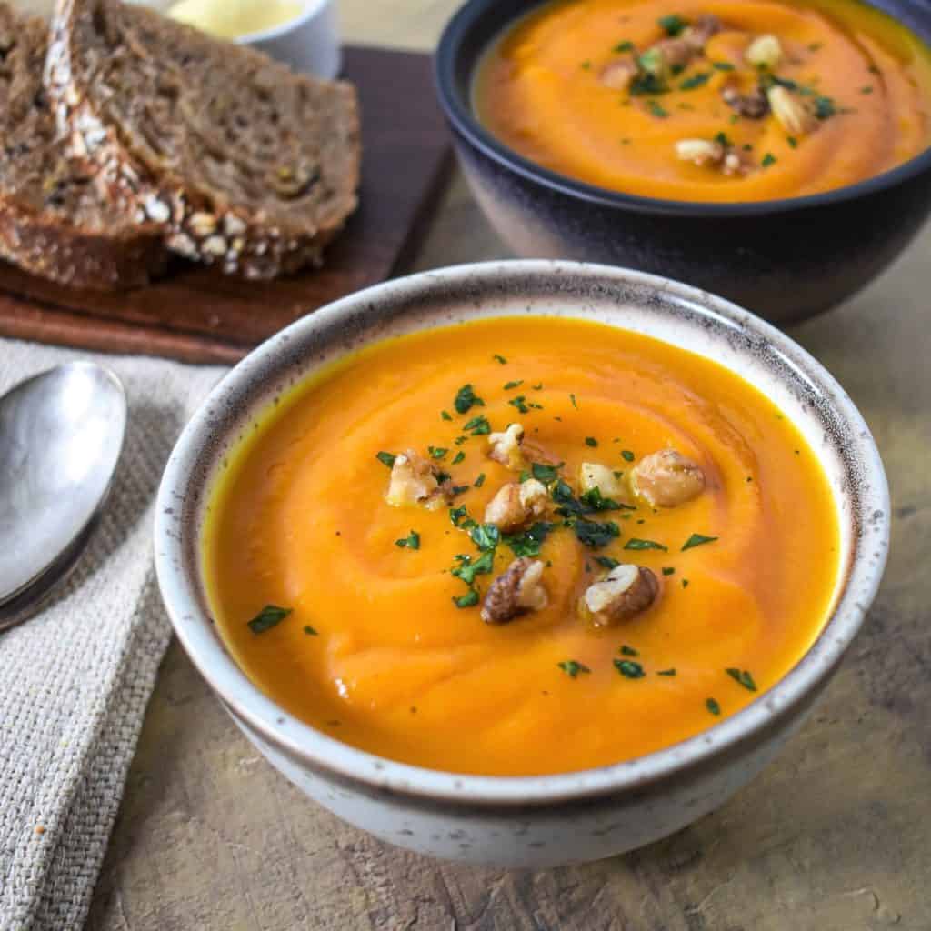 Two carrot sweet potato soups served in small bowls and garnished with parsley and chopped walnuts with bread slices in the background.
