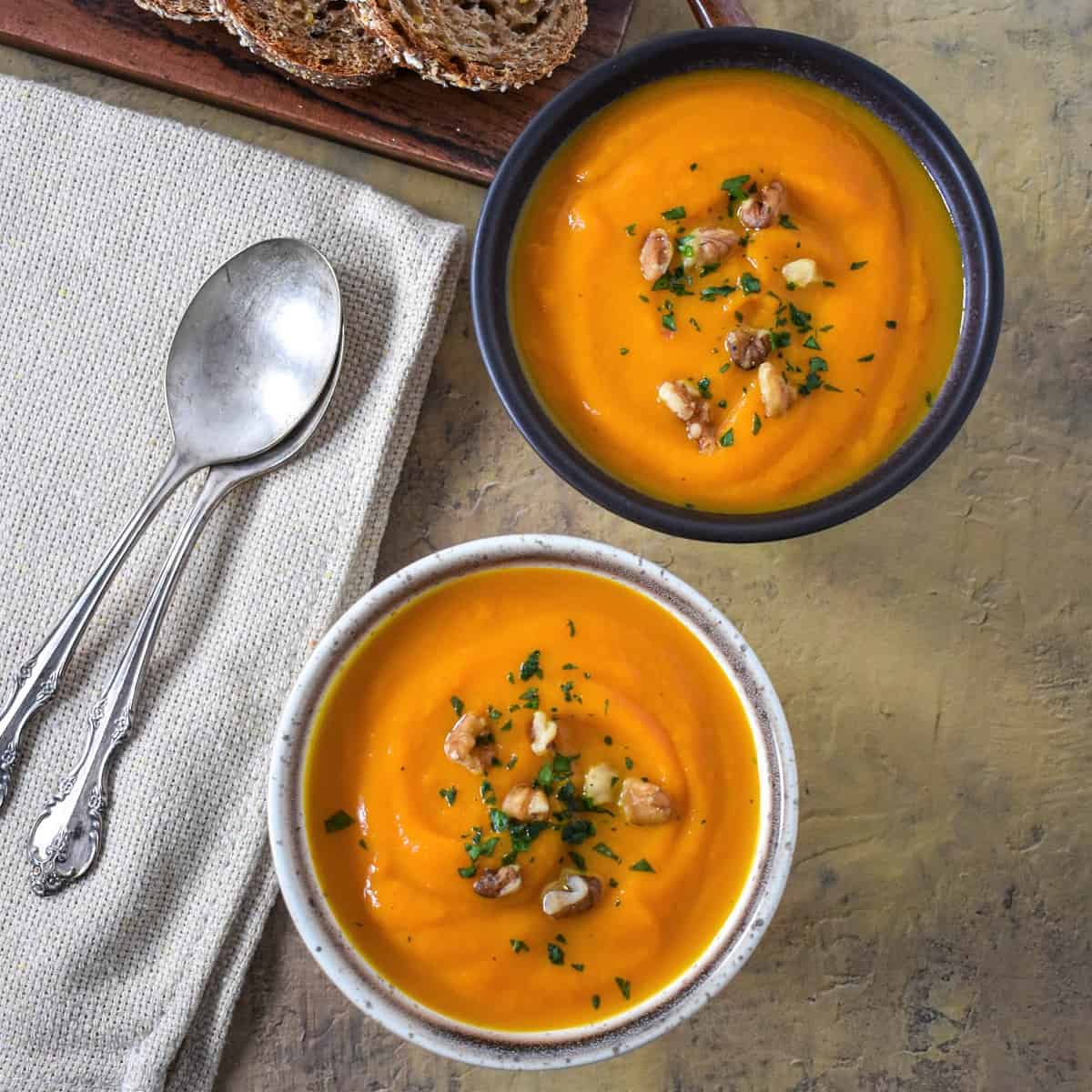 Two carrot sweet potato soups served in small bowls and garnished with parsley and chopped walnuts with bread slices in the background.