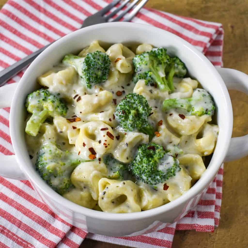 The creamy tortellini with broccoli served in a white bowl set on a red and white striped linen with a fork in the background.