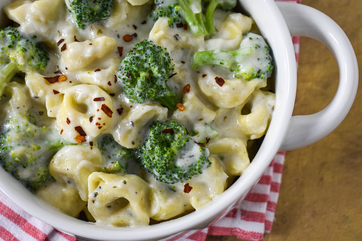 A close up of the creamy tortellini with broccoli served in a white bowl set on a red and white striped linen.