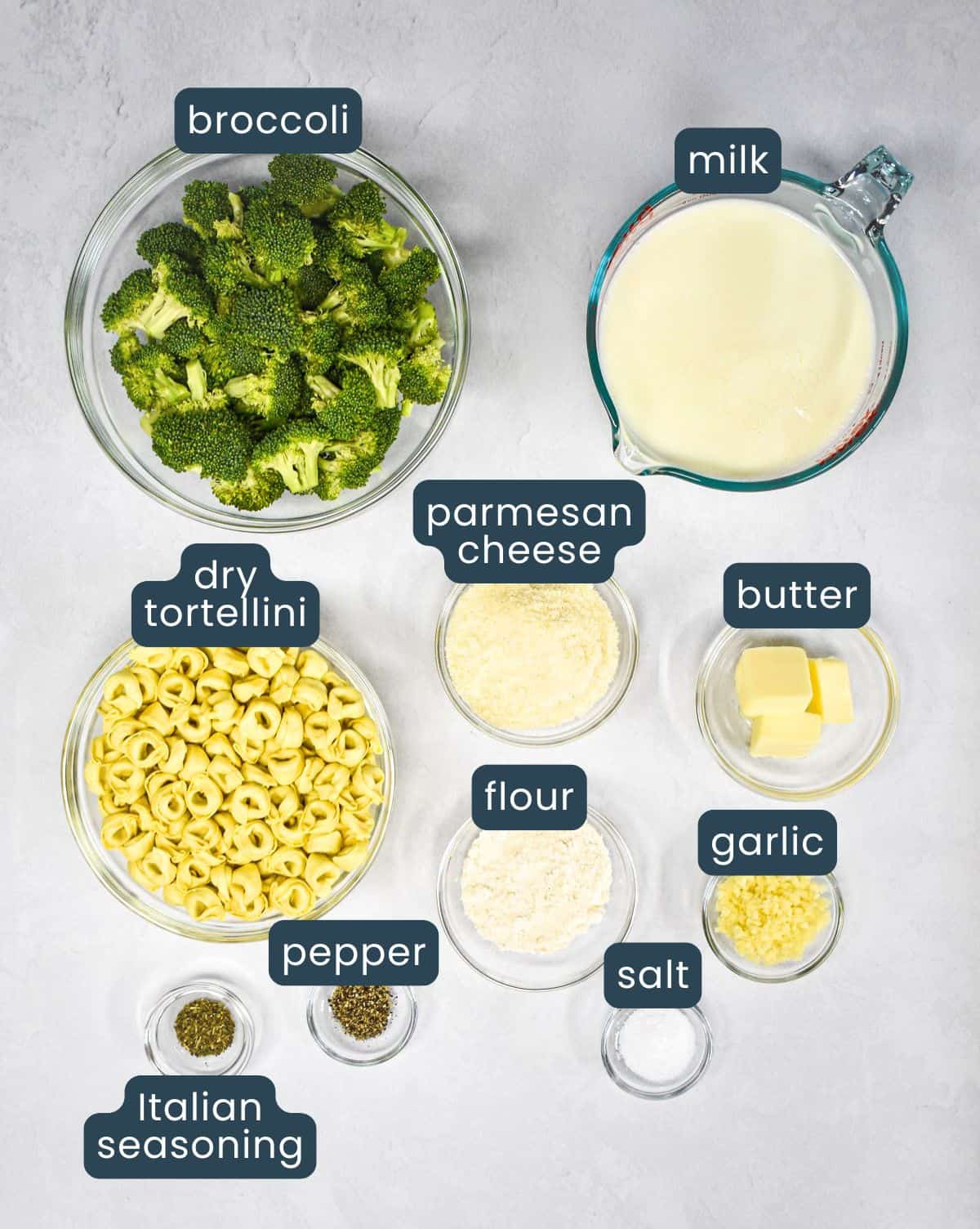 The ingredients for the tortellini with broccoli prepped and arranged in glass bowls set on a light table, with each ingredient labeled.