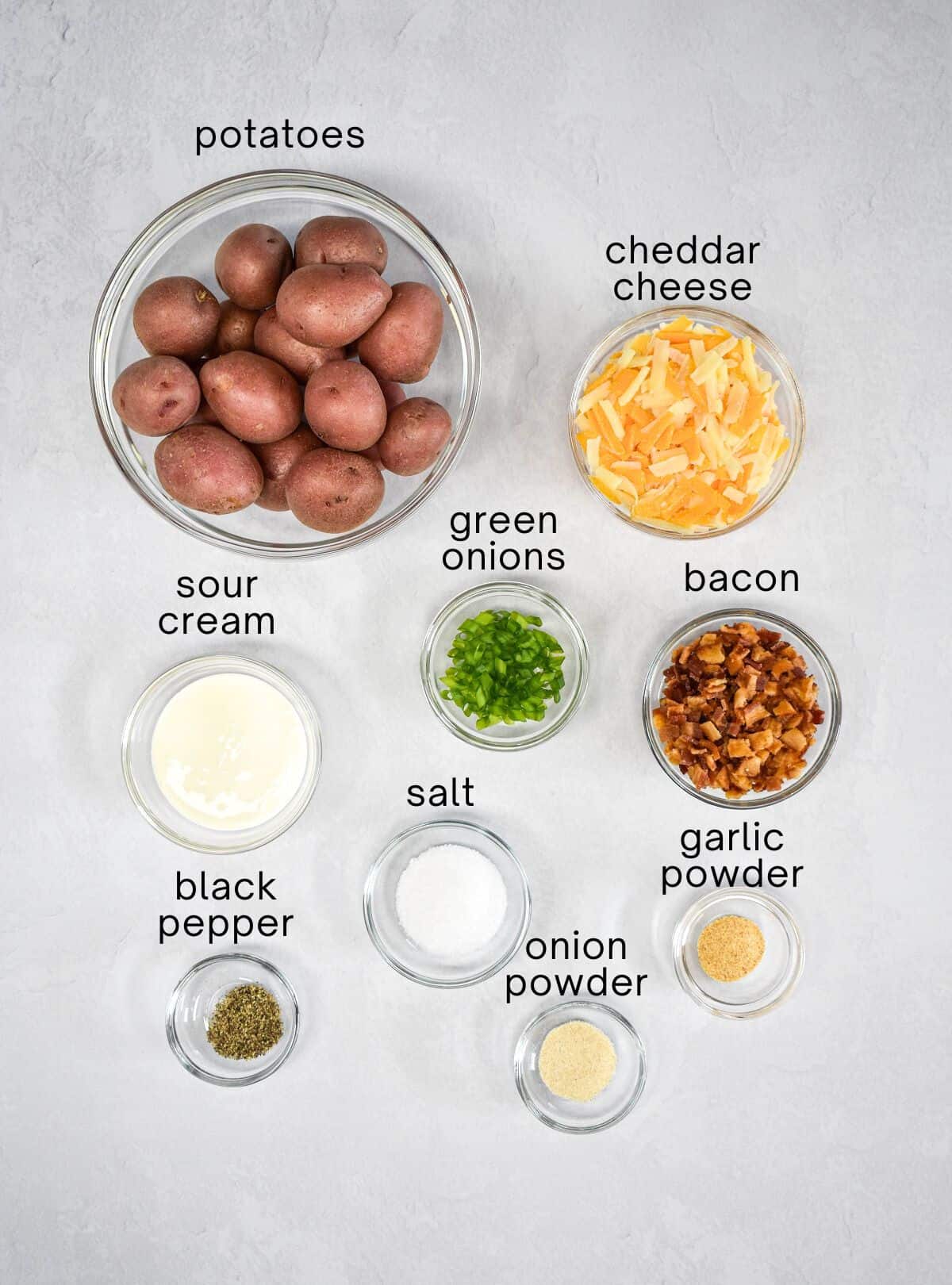The ingredients for the smashed potatoes prepped and arranged in glass bowls on a white table with each labeled with small, black letters.