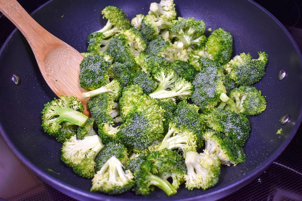 Broccoli florets in a large, black skillet with a wooden spoon to the left side.