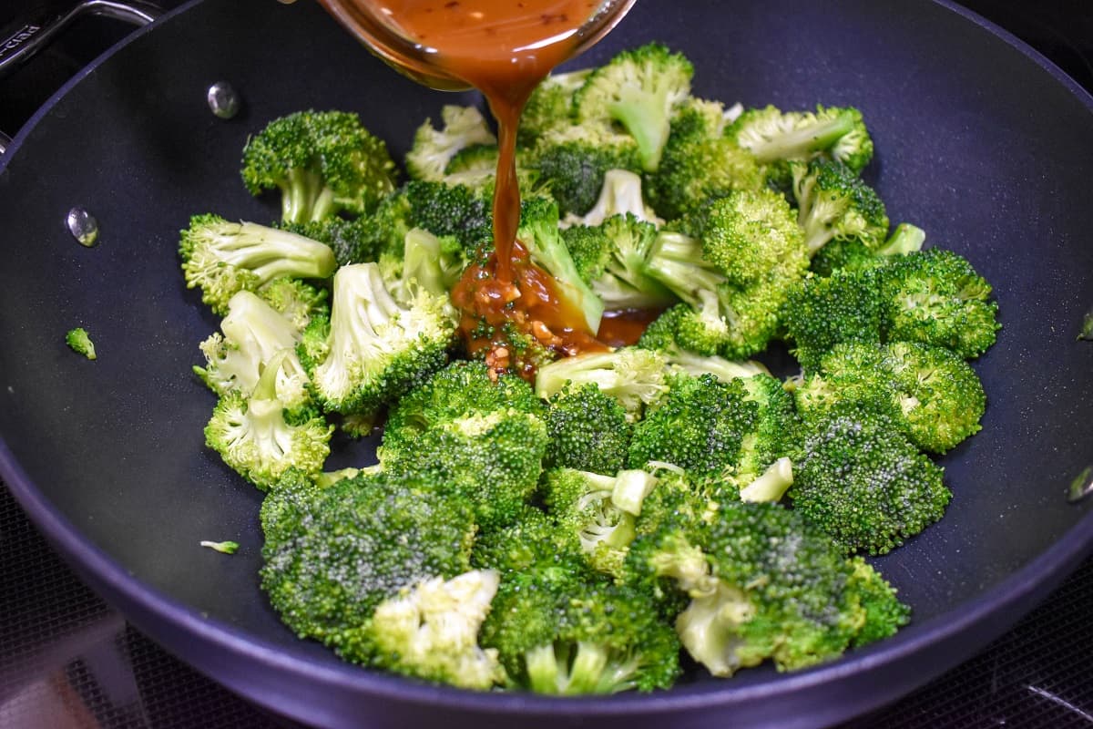 Stir fry sauce being poured over broccoli florets in a large, black skillet.