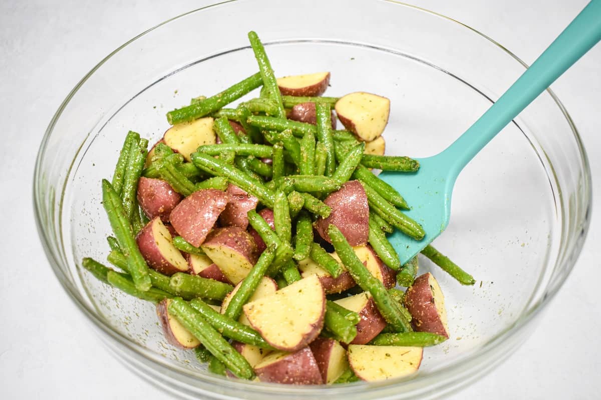 Seasoned green beans and cut potatoes in a large, glass bowl set on a white table.