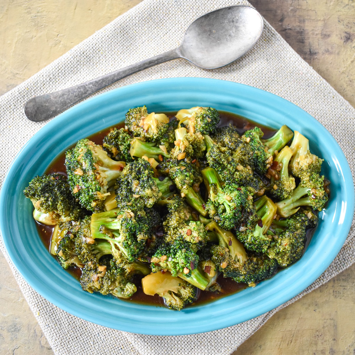 The broccoli stir fry served in an oval, teal plate set on a beige linen with a silver serving spoon on the top, left side.