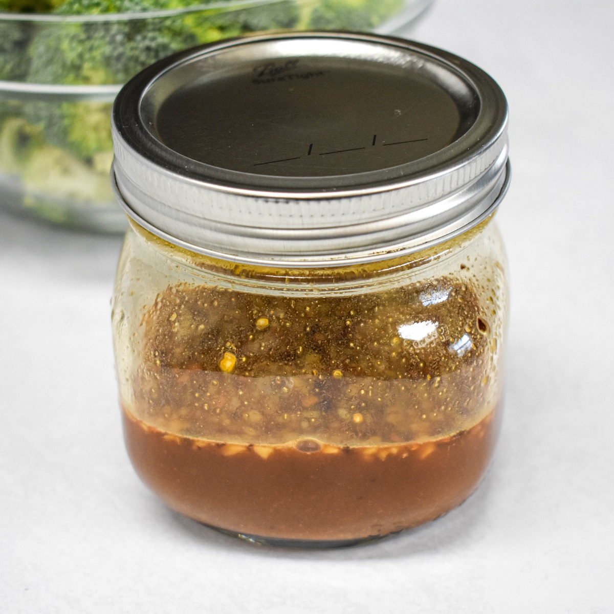 The stir fry sauce in a small mason jar with a bowl of broccoli in the background all set on a white table.
