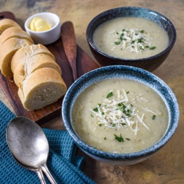 Two bowls of the creamy artichoke soup garnished with parsley and parmesan cheese with sliced bread rounds, butter, and two spoons to the lefthand side.