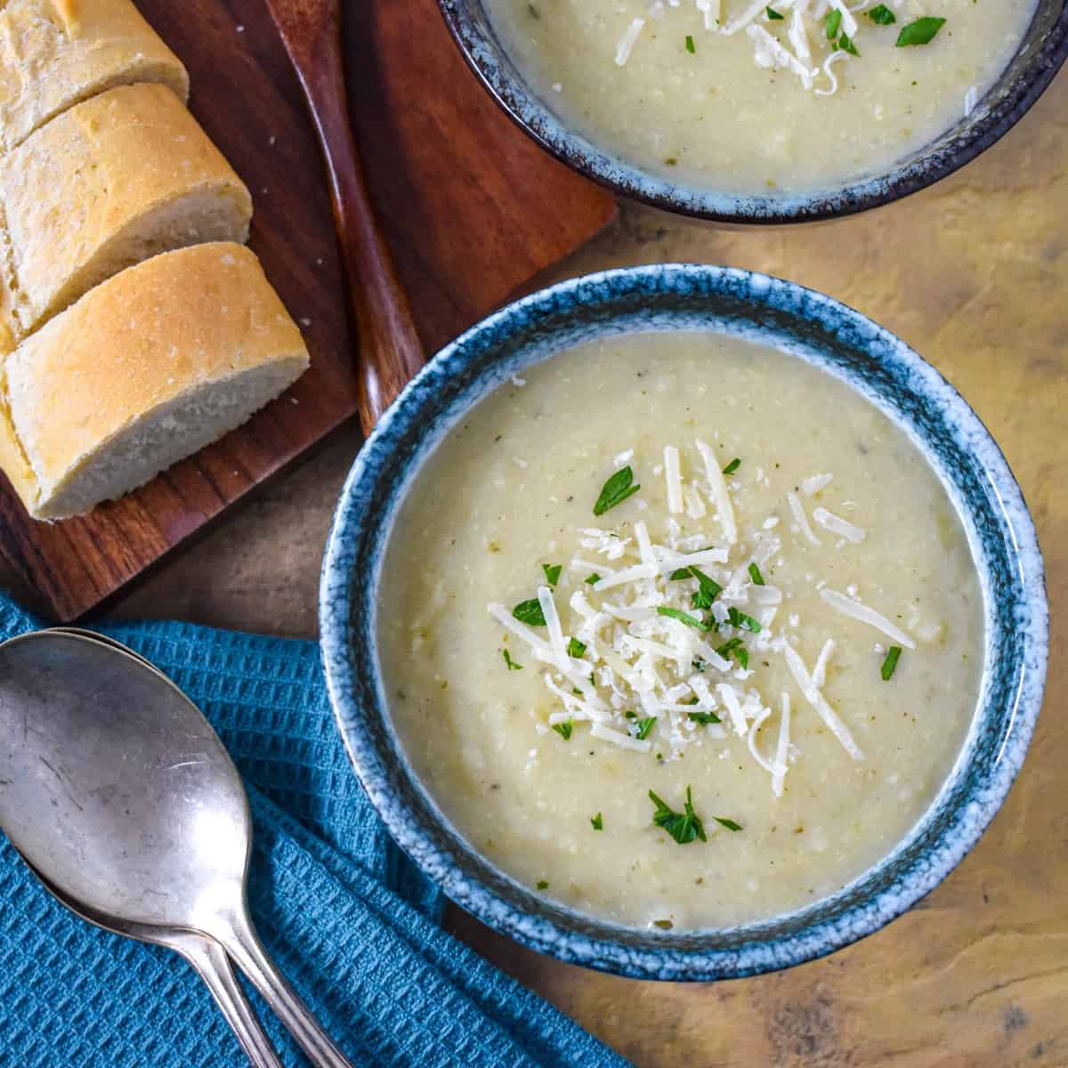 A close-up of the creamy artichoke soup garnished with parsley and parmesan cheese with sliced bread rounds and two spoons to the lefthand side.