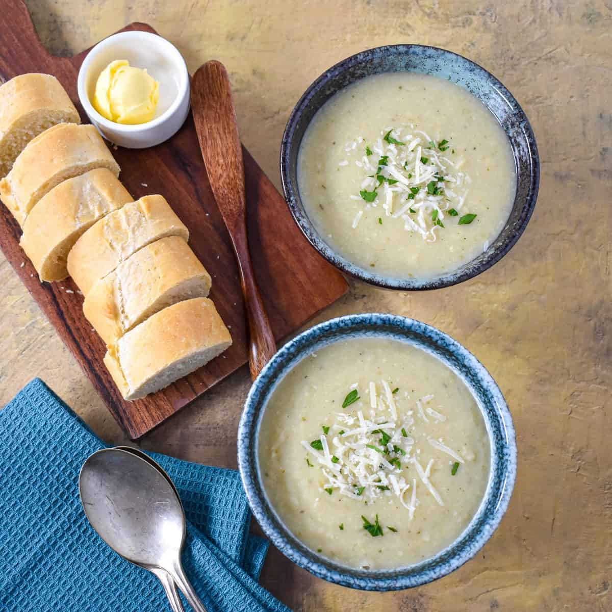 Two bowls of the creamy artichoke soup garnished with parsley and parmesan cheese with sliced bread rounds, butter, and two spoons to the lefthand side.