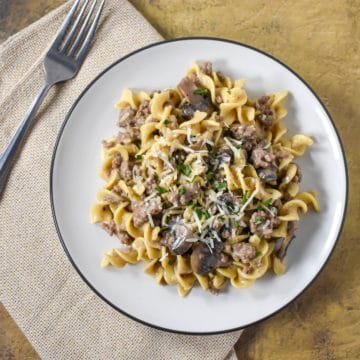 The finished beef stroganoff and egg noodles served on a white plate with a beige linen and fork to the left side.