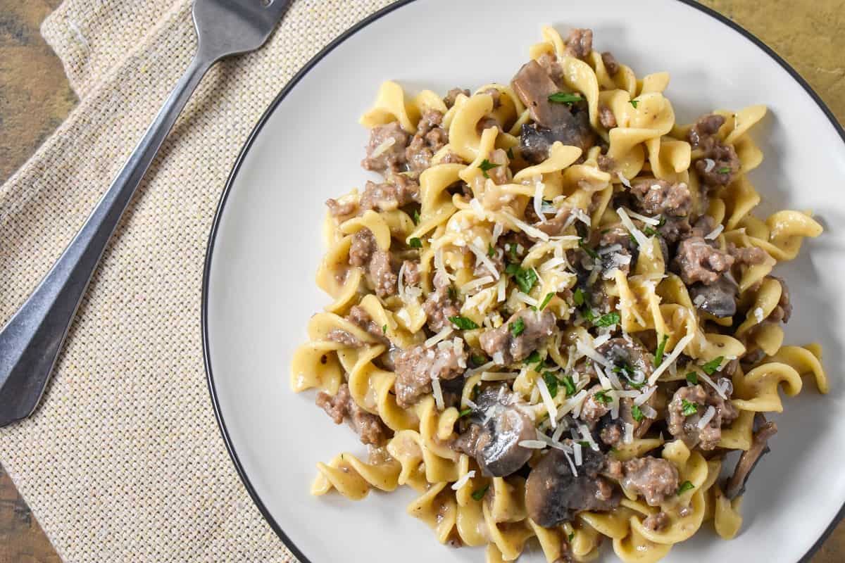 The finished beef stroganoff and egg noodles served on a white plate with a beige linen and fork to the left side.