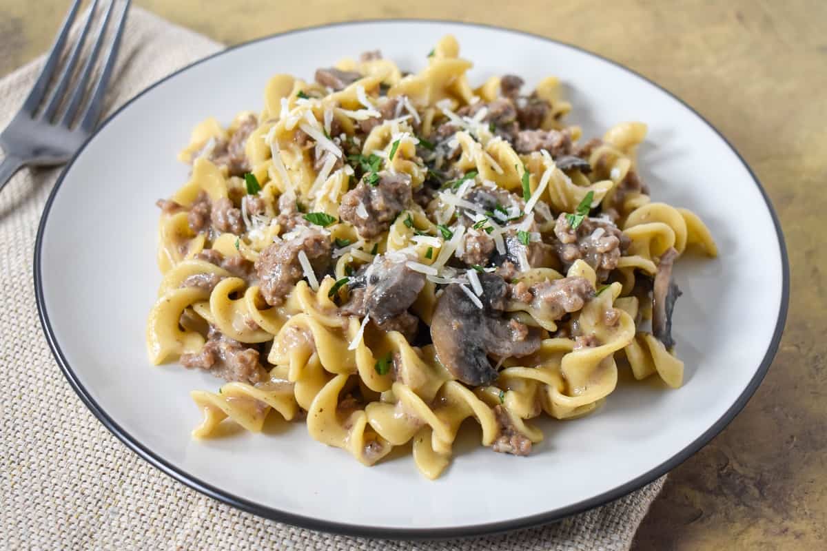 The finished beef stroganoff and egg noodles served on a white plate with a beige linen and fork to the left side.