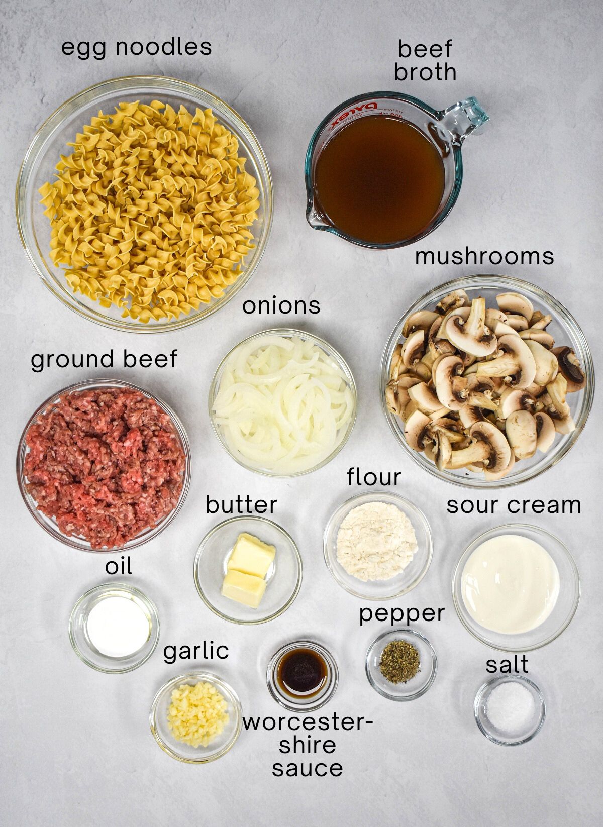 The ingredients for the stroganoff prepped and arranged in glass bowls and set on a white table with each labeled with small, black letters.