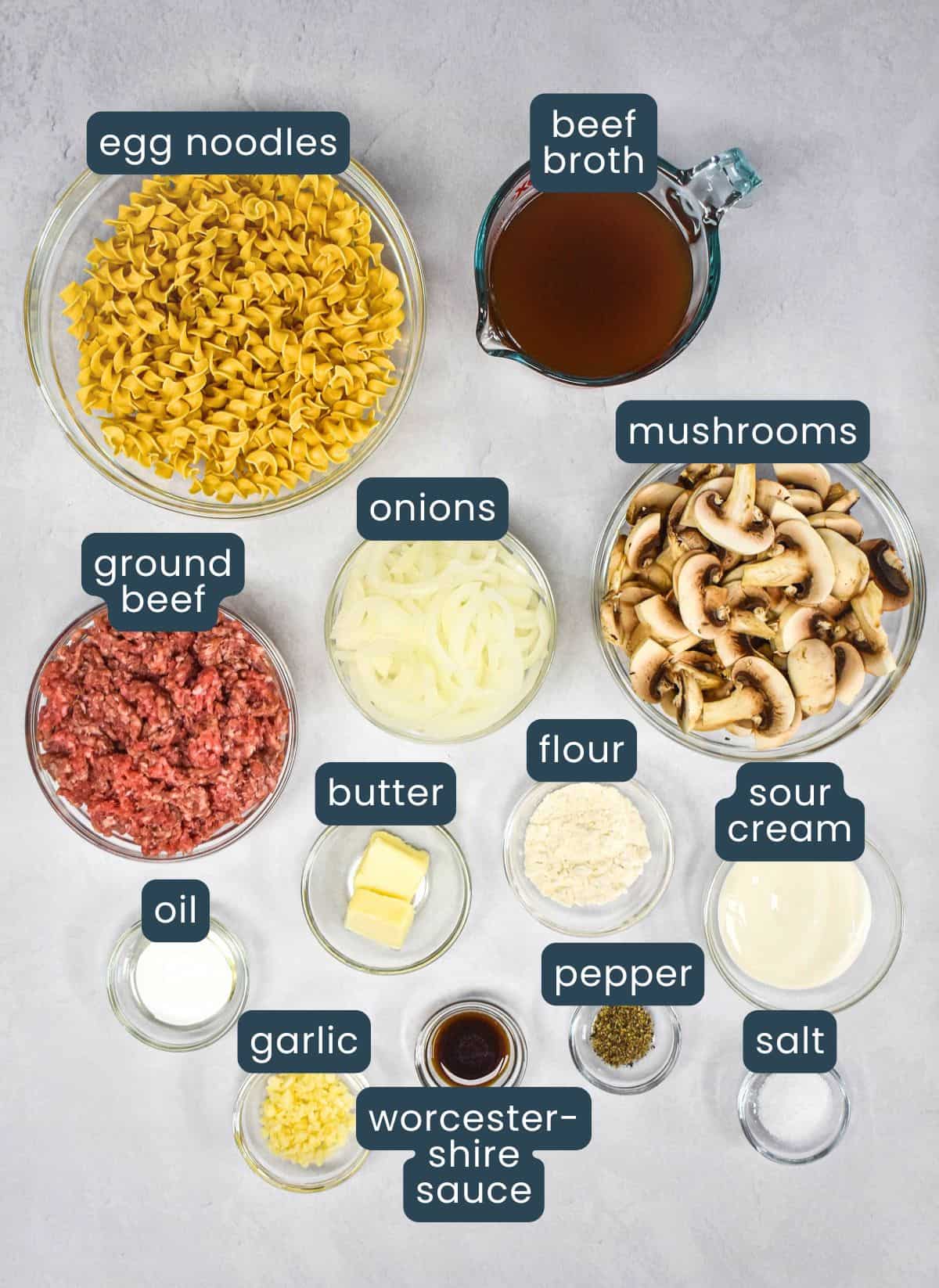 Ingredients for ground beef stroganoff in glass bowls on a white table, labeled egg noodles, beef broth, mushrooms, onions, ground beef, butter, flour, sour cream, oil, garlic, Worcestershire sauce, pepper, and salt.