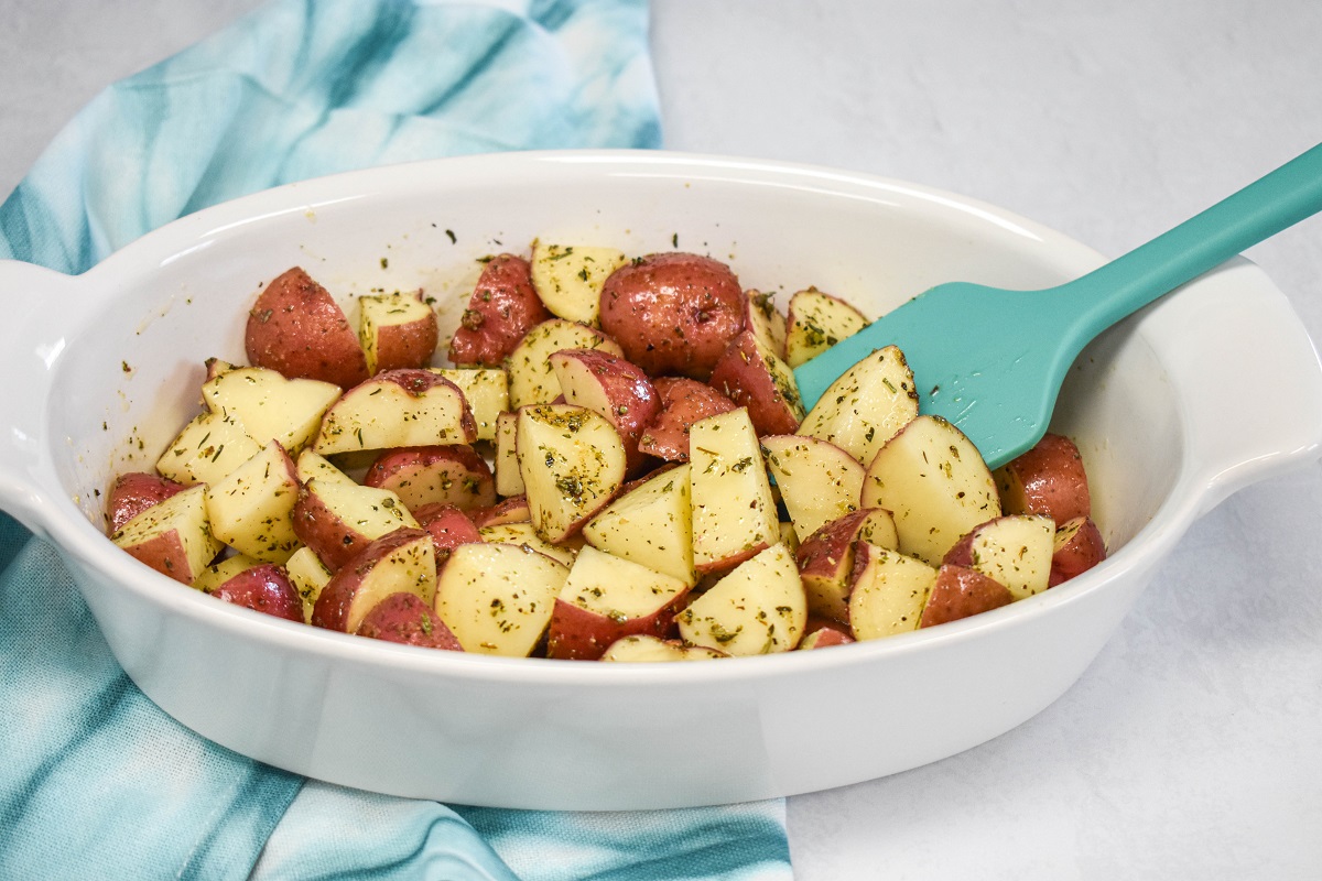 Seasoned cut potatoes in a white baking dish set on a white talbe with a teal linen and a silicone spatual to the right.