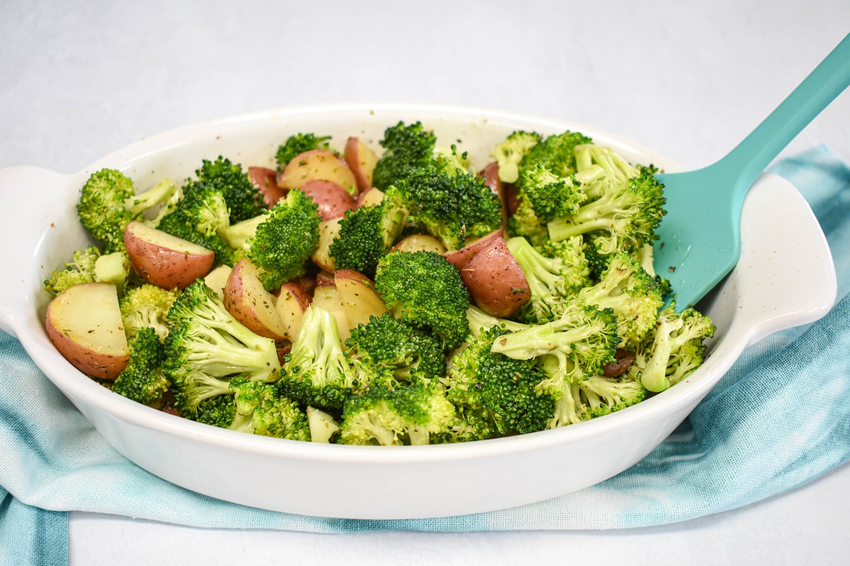 Broccoli florets and cut red potatoes in a white baking dish set on a teal linen with a silicone spatula to the right.