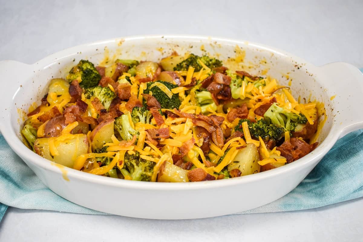 Baked broccoli and potatoes covered with cheese and bacon in a white casserole dish set on a teal linen on a white table.