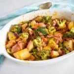 The potato broccoli bake in a white casserole dish set on a white table with a teal and white linen and a serving spoon.