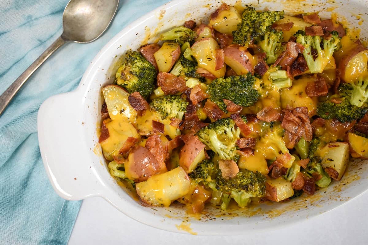 The potato broccoli bake in a white casserole dish set on a white table with a teal linen and a serving spoon.