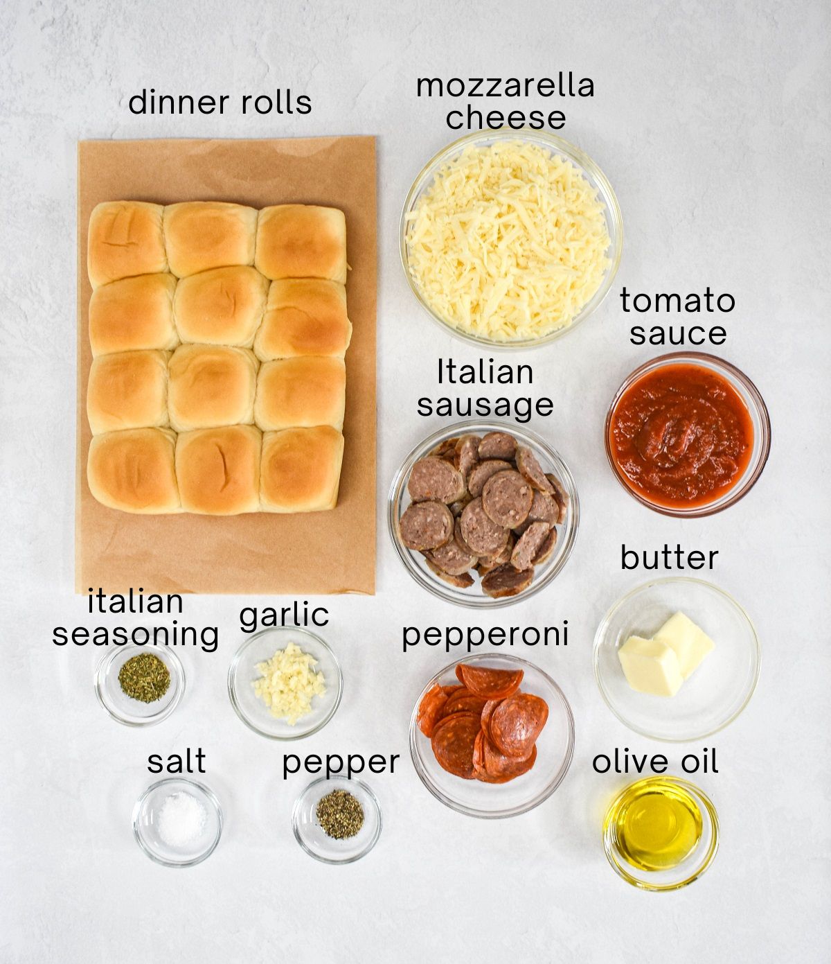 The ingredients for the pepperoni sausage pizza sliders prepped and arranged in glass bowls on a white table with each one labled with small, black letters.
