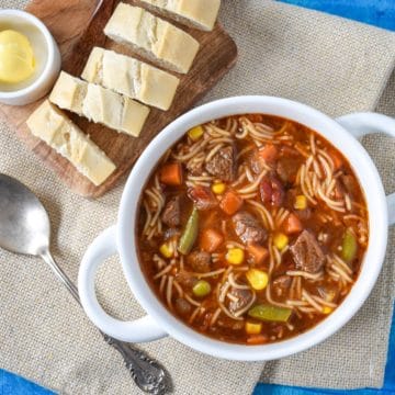 The beef soup with noodles served in a white bowl with sliced bread and butter on a small cutting board in the back.