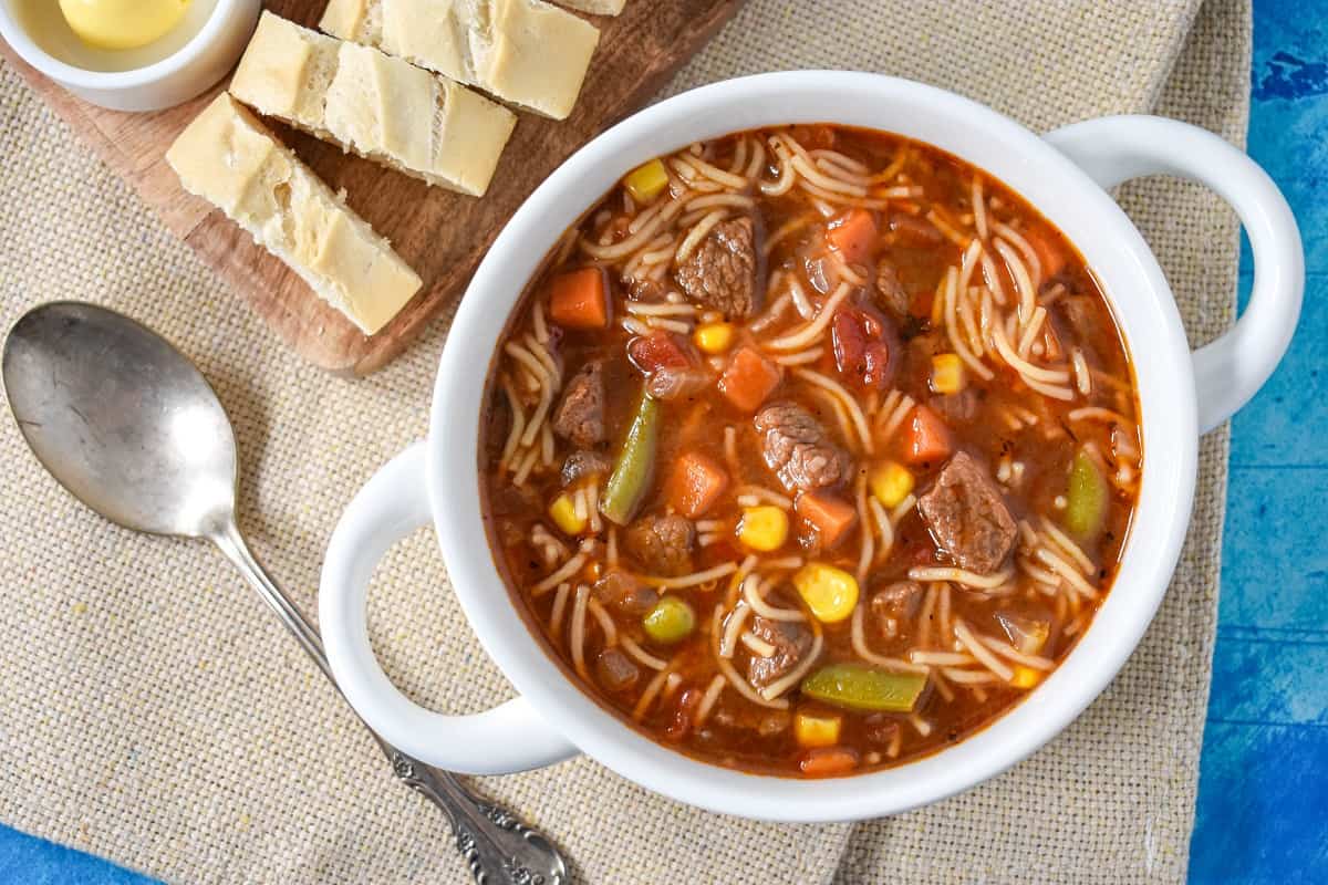 The beef soup with noodles served in a white bowl with sliced bread and butter on a small cutting board in the back.