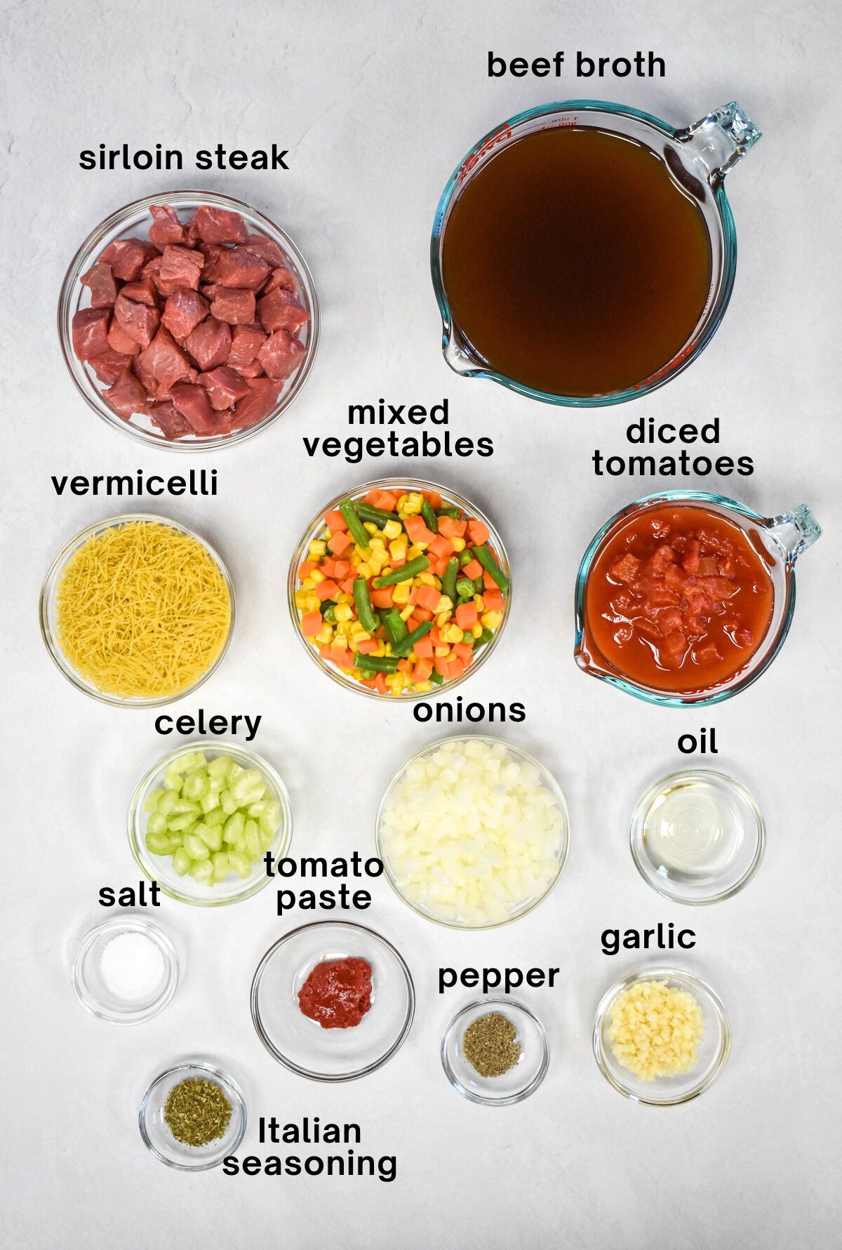 The ingredients for the soup prepped and arranged in glass bowls set on a white table with each labeled with small black letters.