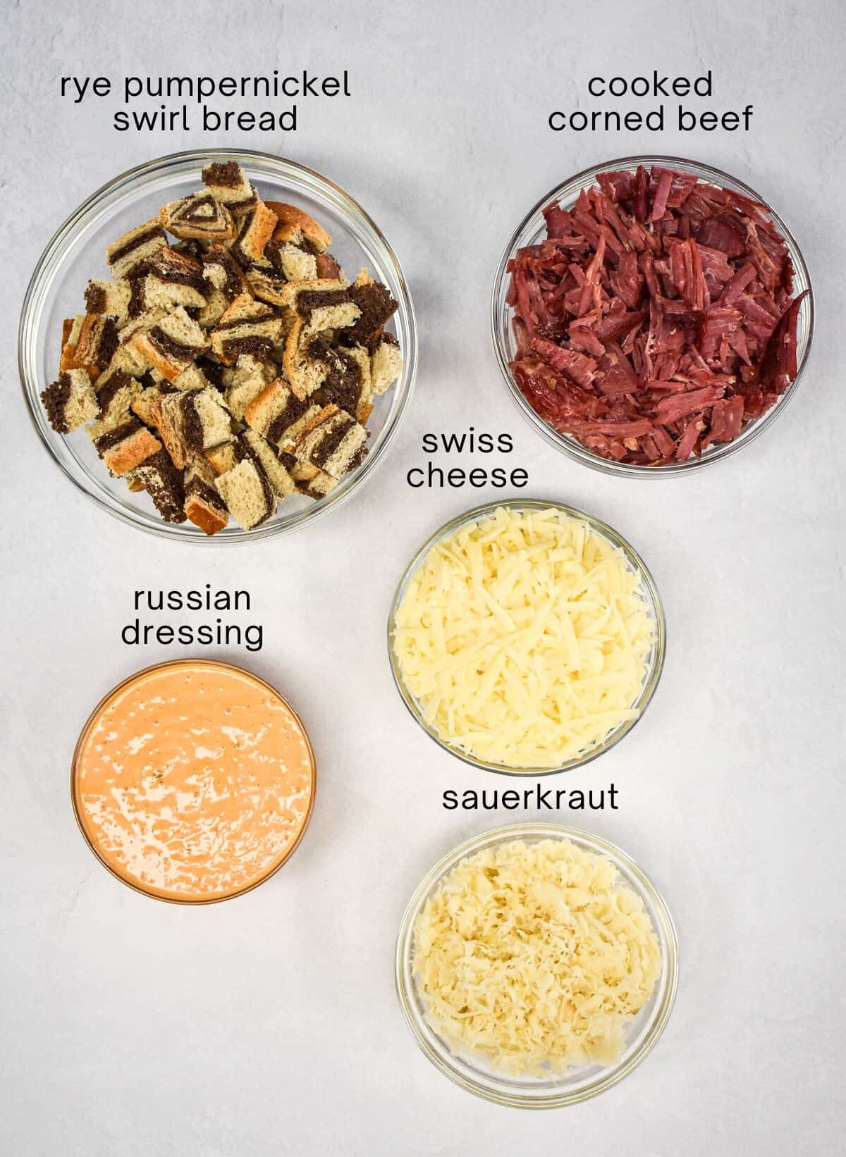 The ingredients for the casserole prepped and arranged in glass bowls on a white table with each labeled in small, black letters.