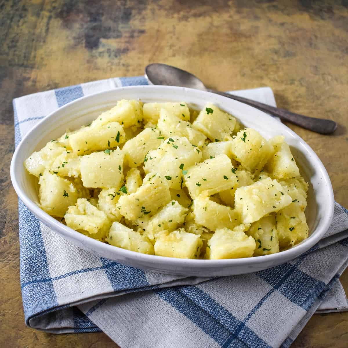 The yuca with garlic sauce served in an oval white bowl set on a blue and white linen with a serving spoon.