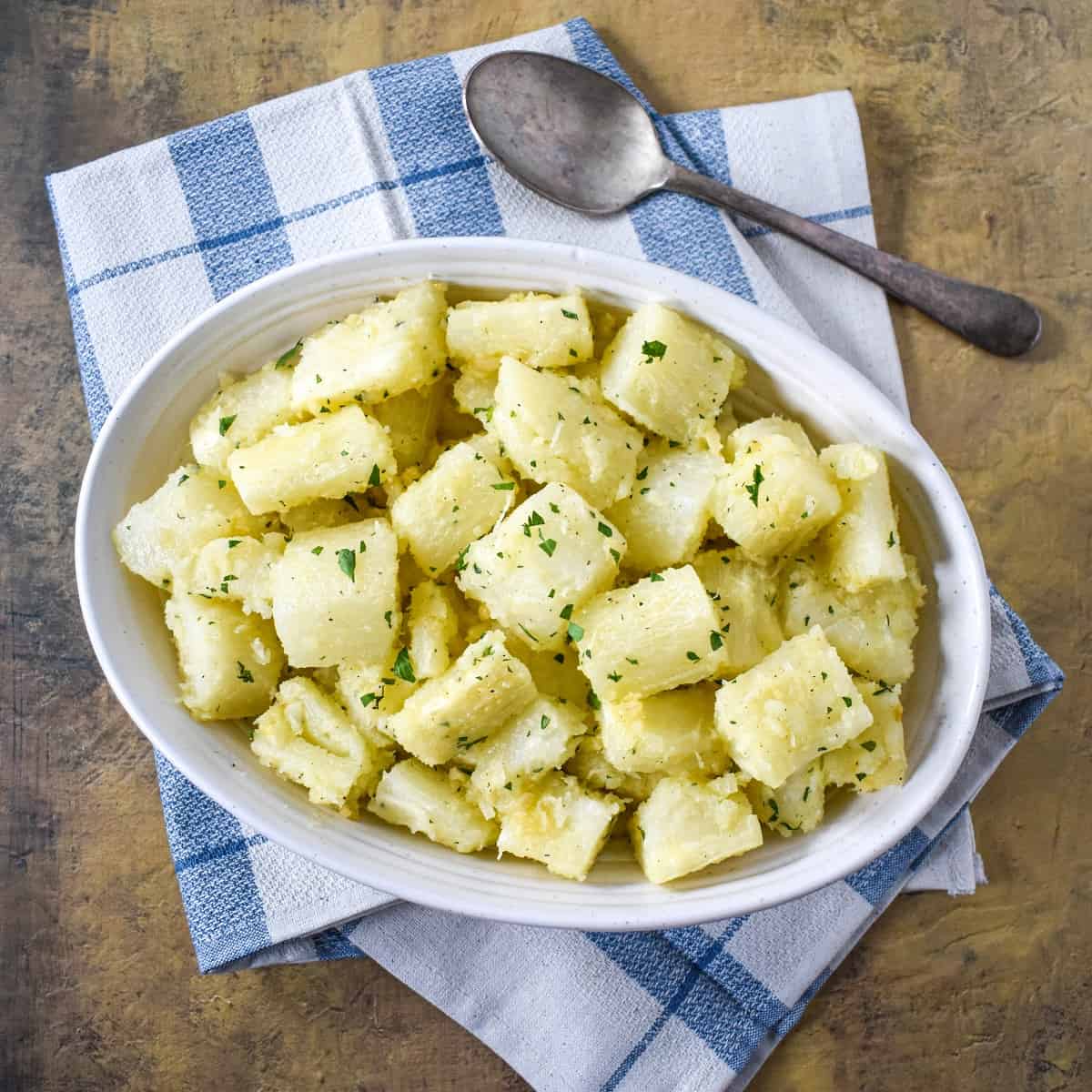 The yuca with garlic sauce served in an oval white bowl set on a blue and white linen with a serving spoon.