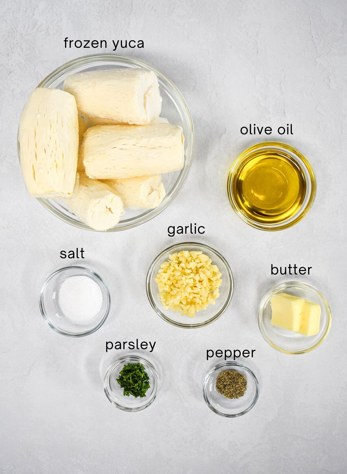 The ingredients for the dish prepped in glass bowls set on a white table with each one labeled with small, black letters.