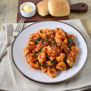 The Italian sausage tortellini served on a white plate, set on a beige linen with bread rolls and butter in the background.