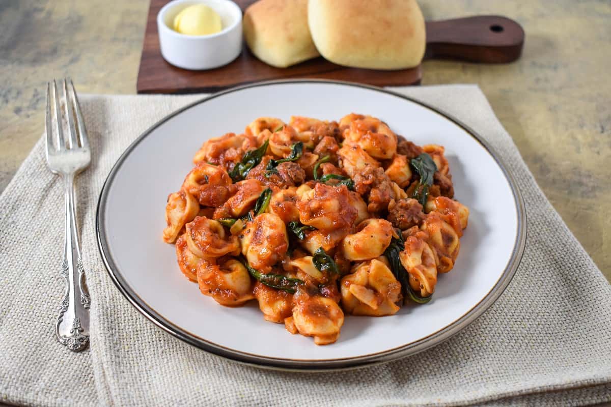 The Italian sausage tortellini served on a white plate, set on a beige linen with bread rolls and butter in the background.