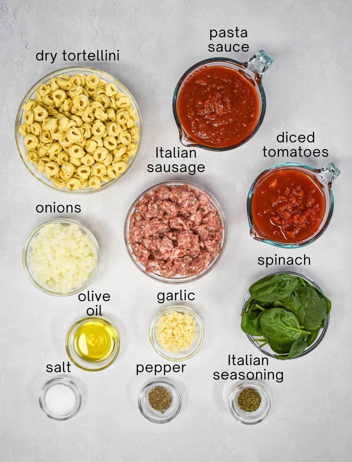 The ingredients for the pasta dish prepped and arranged in glass bowls on a white table with each one labeled with small, black letters.