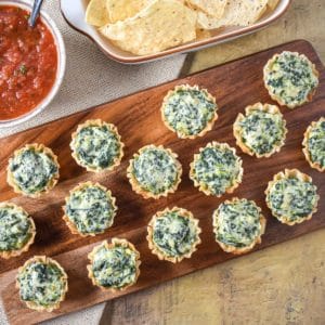 The spinach artichoke bites served on a wood board with salsa and chips in the background.