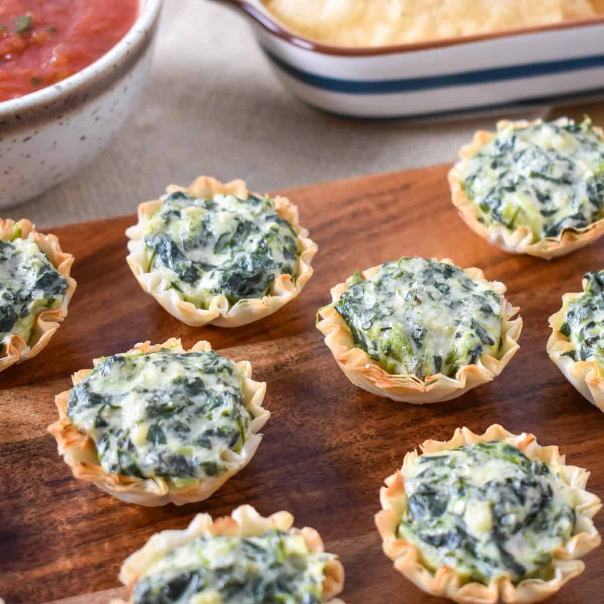 The spinach artichoke bites served on a wood board with salsa and chips in the background.