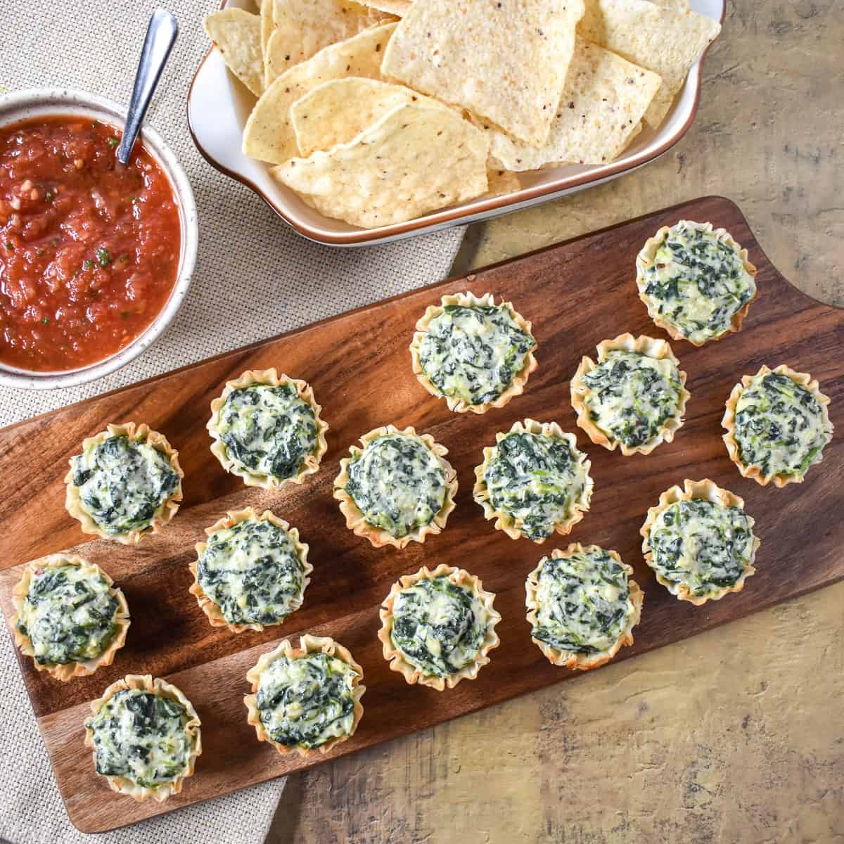 The spinach artichoke bites served on a wood board with salsa and chips in the background.