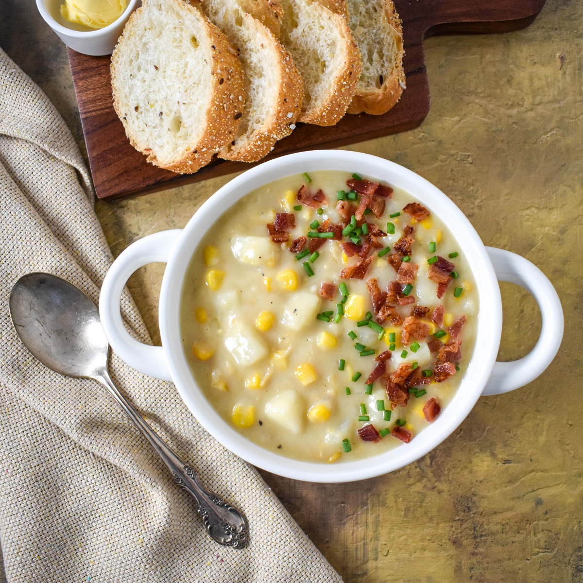 The potato corn chowder garnished with bacon bits and chives served in a white bowl with sliced bread on a board in the background.
