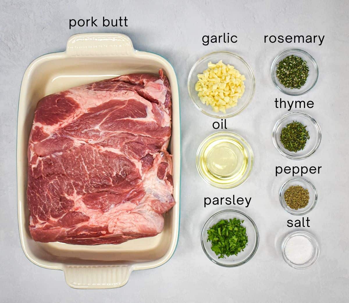 The ingredients for the easy oven roasted pork butt with garlic and herbs prepped and arranged on a white table with each one labeled with small, black letters.