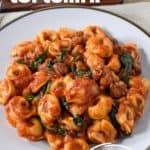 The sausage tortellini served on a white plate, set on a beige linen with bread rolls and butter in the background.