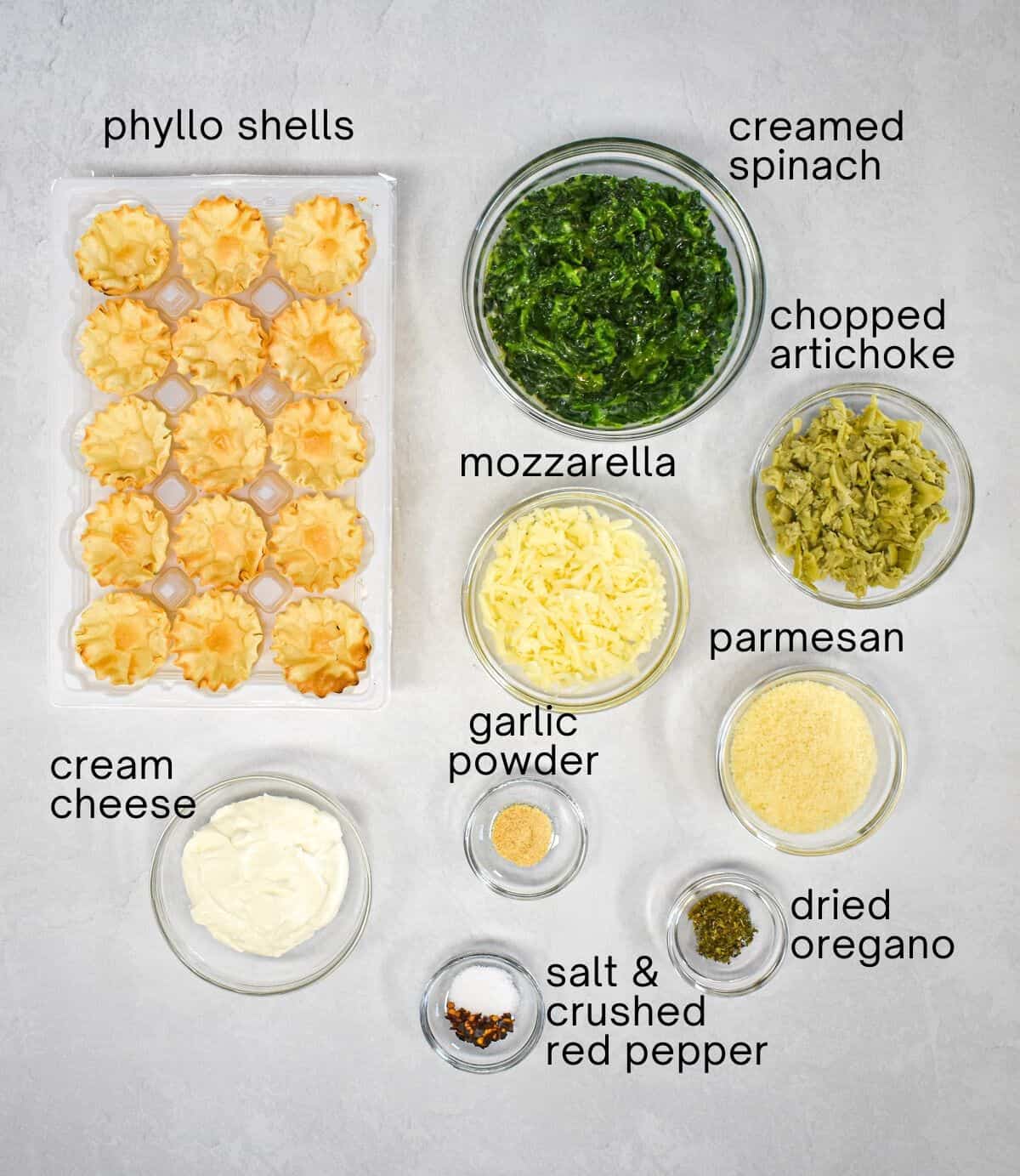 The ingredients for the spinach artichoke cups arranged on a white table with each labeled with small black letters.