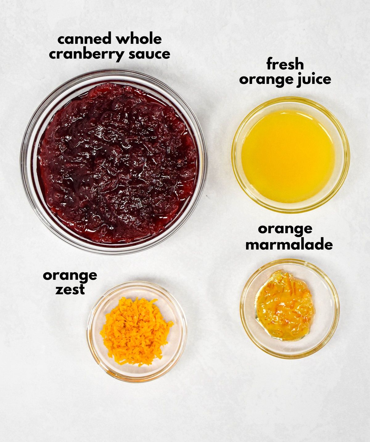 The ingredients for the cranberry sauce arranged in glass bowls on a white table with each labeled with small, black letters.
