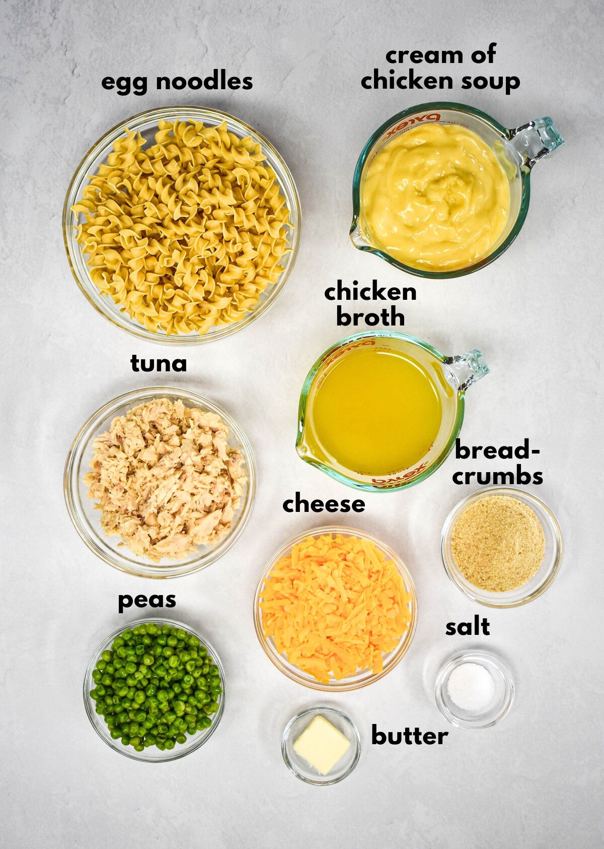 The ingredients for the casserole arranged in glass bowls on a white table with each labeled with small black letters.