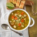 The turkey barley soup served in a white bowl set on a linen with sliced bread and lime wedges in the background.