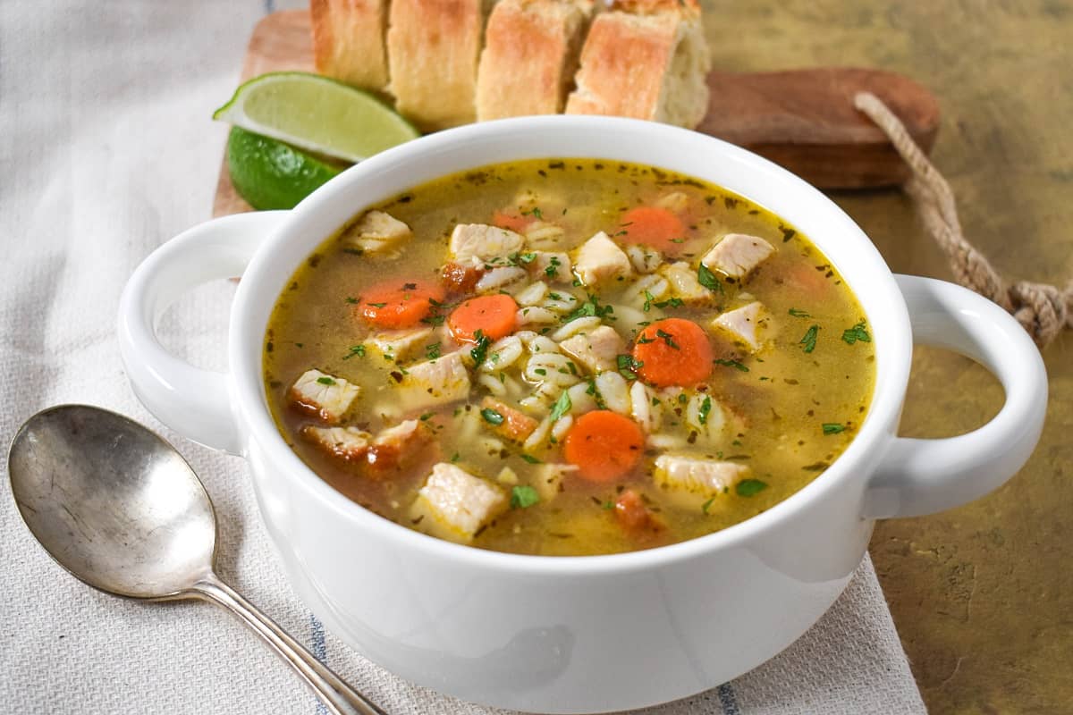 The finished turkey barley soup, garnished with chopped parsley, served in a white bowl set on a linen with sliced bread and lime wedges in the background.