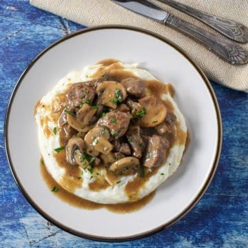 The beef tips and mushrooms served on a bed of mashed potatoes on a white plate.