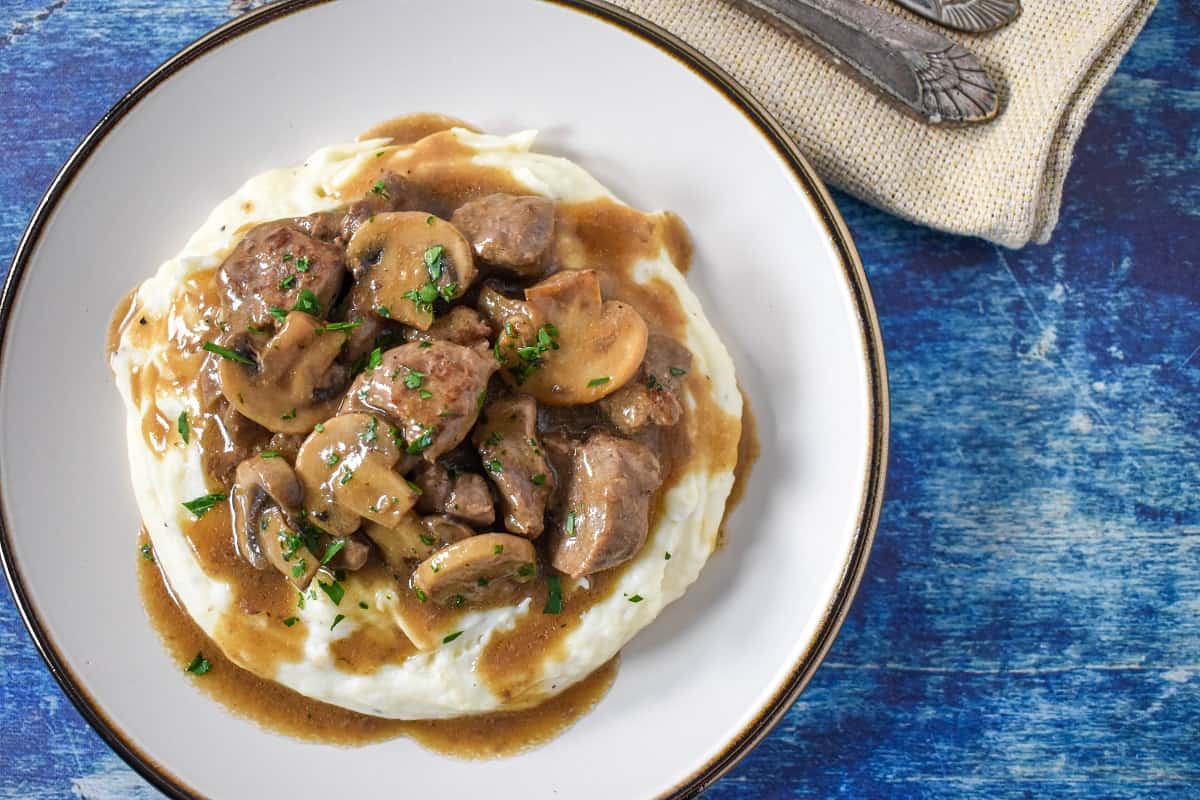 The beef tips and mushrooms served on a bed of mashed potatoes on a white plate.