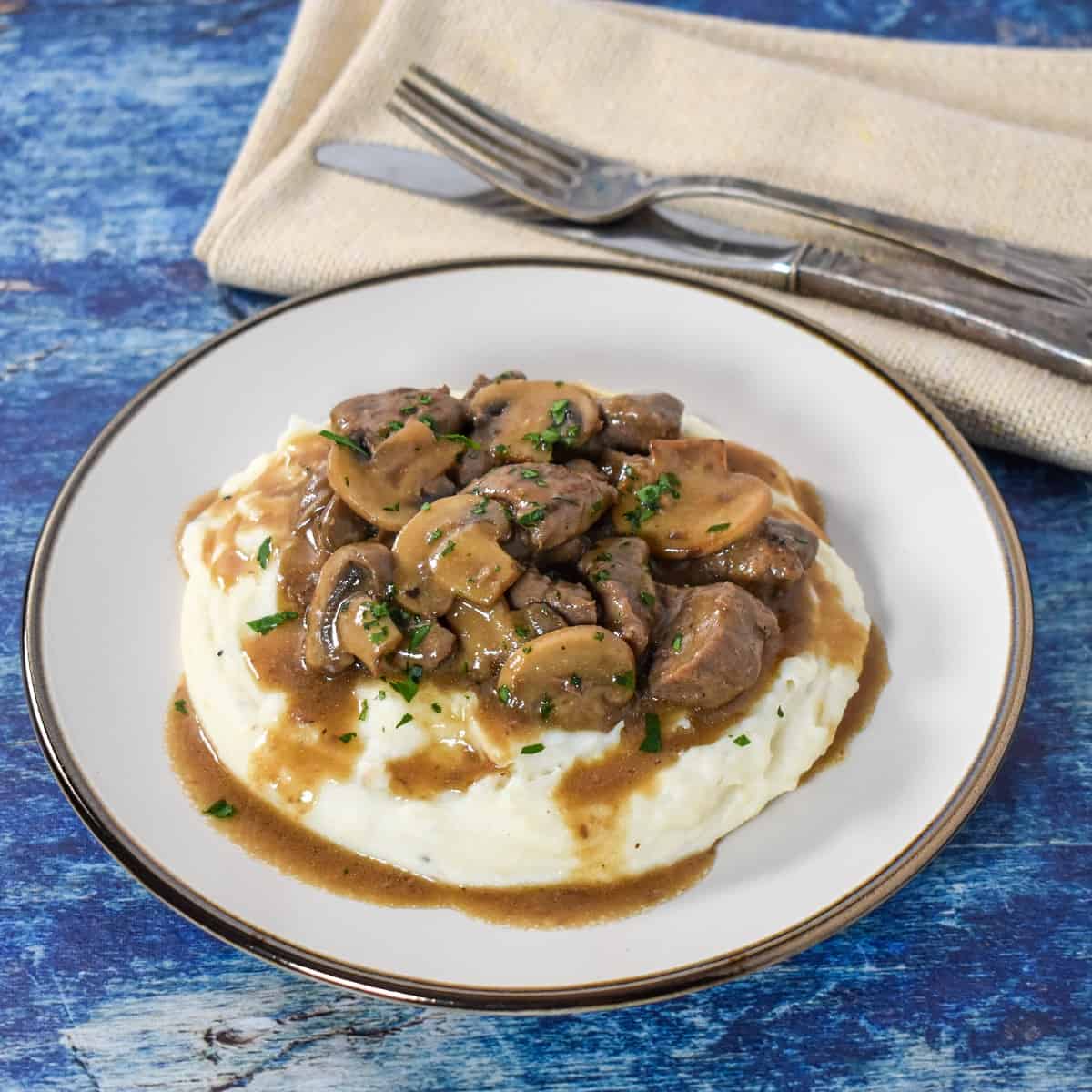 The beef tips and mushrooms served on a bed of mashed potatoes on a white plate with a beige linen and fork and knife in the background.